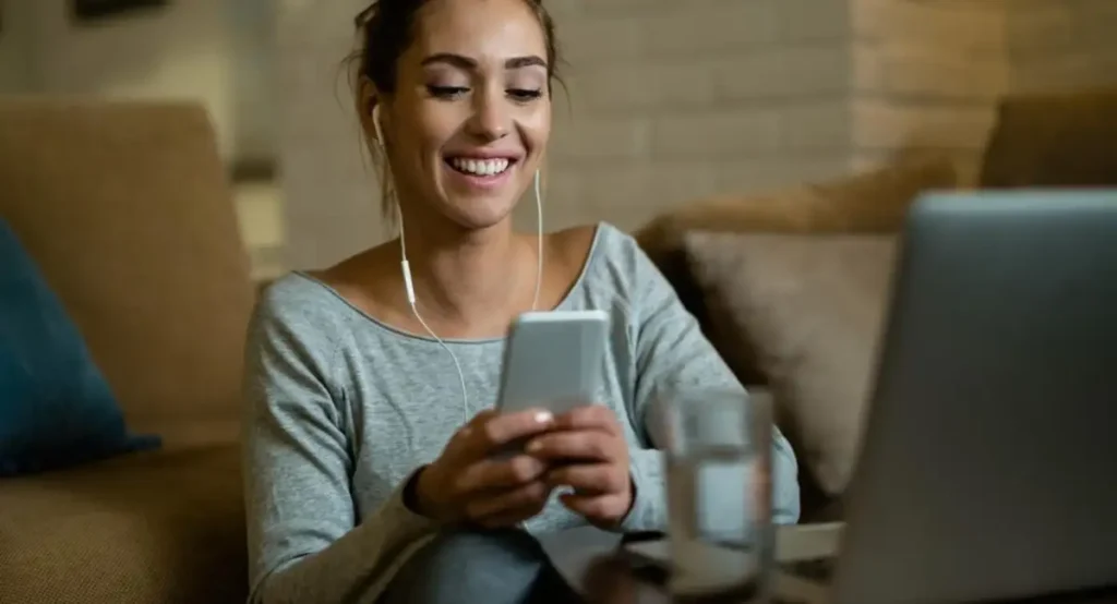 Mulher sorrindo enquanto usa o smartphone e um laptop.