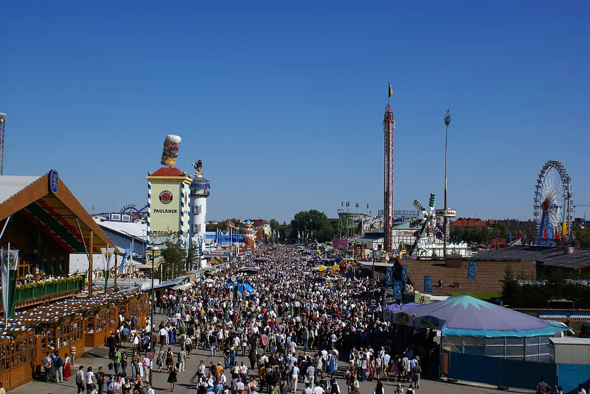 Vista panorâmica da Oktoberfest, com multidão reunida ao redor das tendas, brinquedos e decorada com elementos tradicionais alemães.