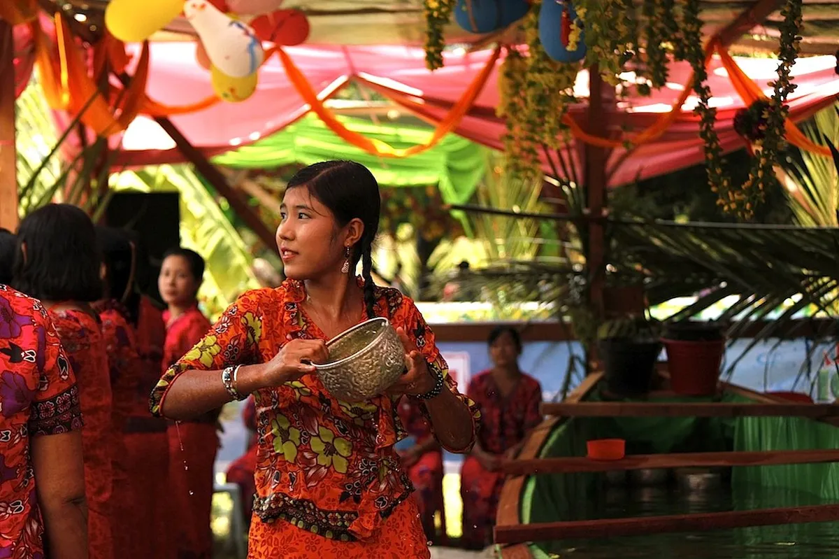 Mulher participando do festival Songkran, com vestimentas tradicionais tailandesas, segurando um recipiente de água durante a celebração.