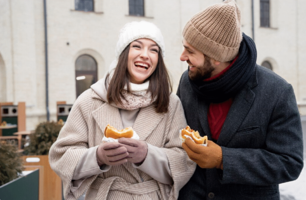 Casal sorridente aproveitando o frio comendo lanche ao ar livre, explorando o aumento do apetite no inverno.