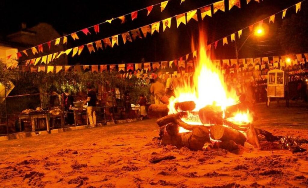 Fogueira acesa em festa junina com bandeirinhas coloridas e pessoas ao fundo