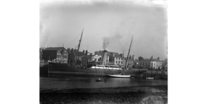Imagem em preto e branco do navio ancorado no porto com edifícios históricos ao fundo, na costa e com farol auxiliar 'hesperus'.