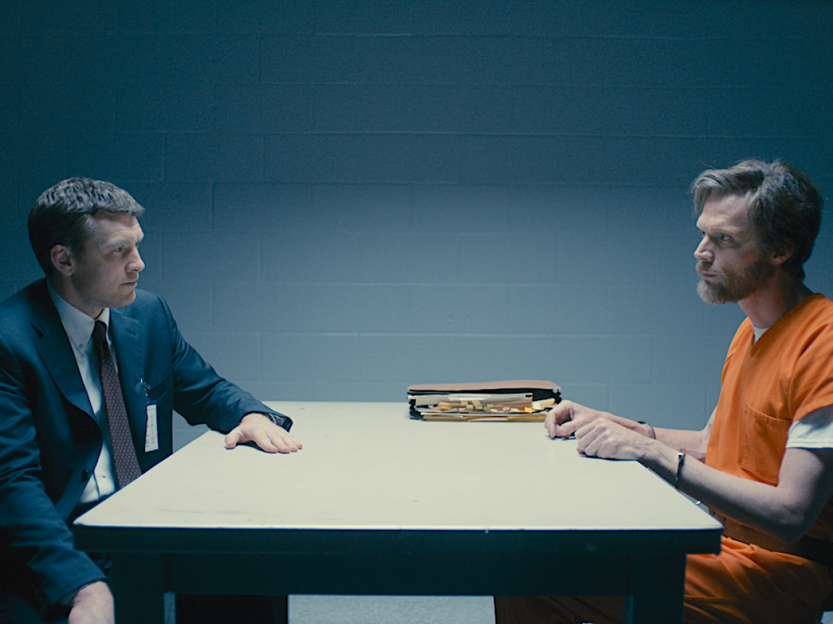 Cena de interrogatório em uma sala de polícia, com um investigador e um prisioneiro em trajes laranja, com papéis sobre a mesa, da nissérie Manhunt Unabomber.