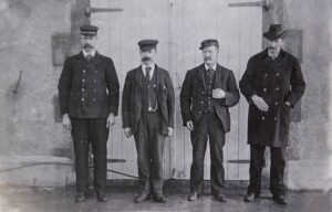 Imagem em preto e branco de quatro homens com bigodes, faloeiros, usando uniformes antigos, posando na frente de uma porta de madeira.